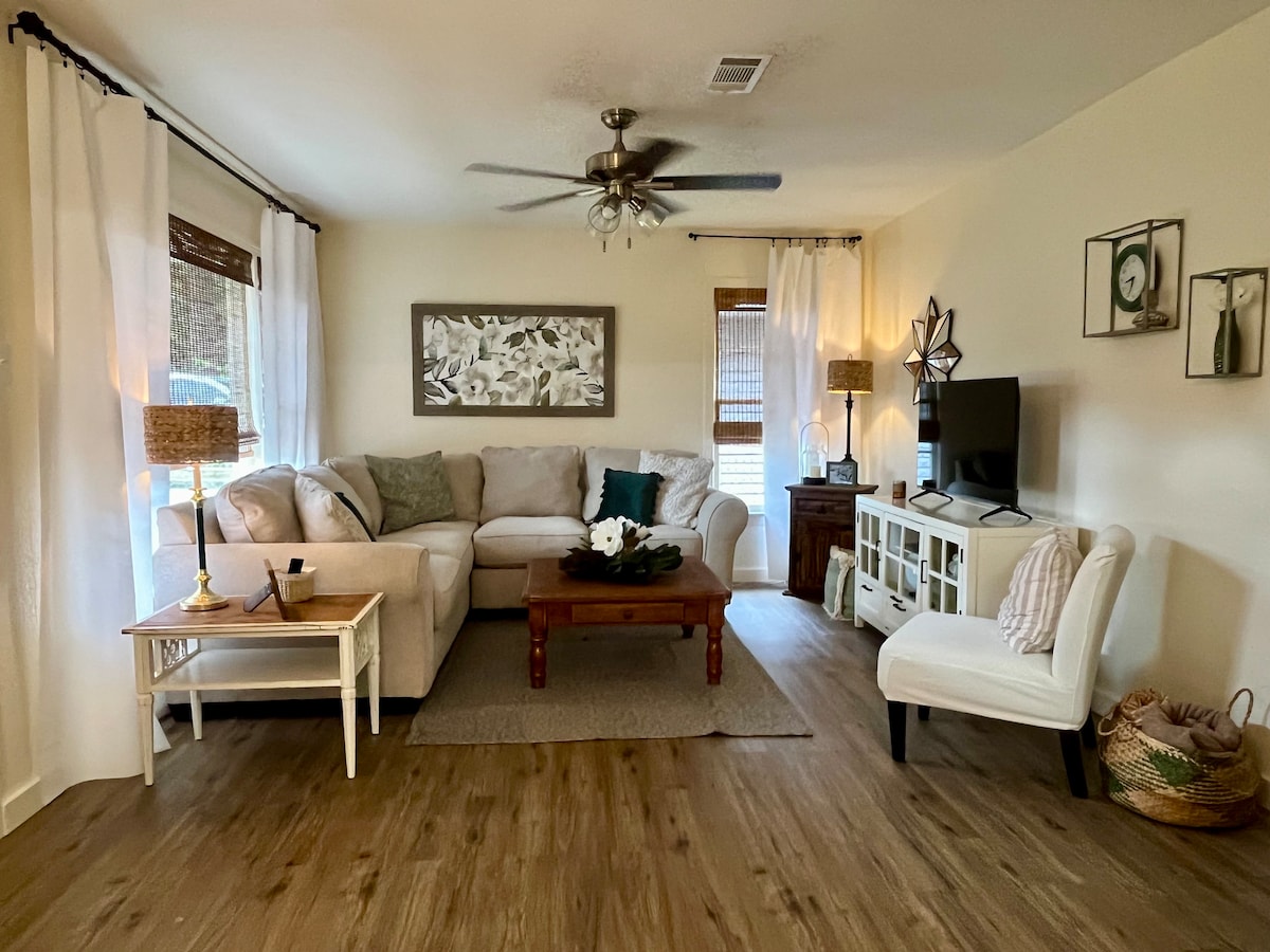 A welcoming living area features a light-colored sectional sofa adorned with assorted cushions. A wooden coffee table is positioned in the center, accompanied by a side table with decorative items. Soft lighting emanates from nearby lamps, enhancing the inviting ambiance.