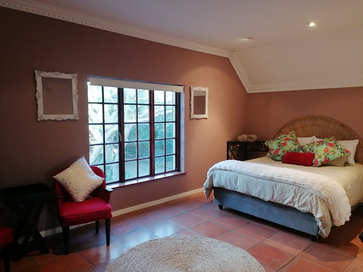A warm-toned room features a spacious bed with decorative pillows and a woven headboard. A small round rug lies on the tiled floor, complemented by two red accent chairs and a side table. Natural light filters through a large window, surrounded by greenery.