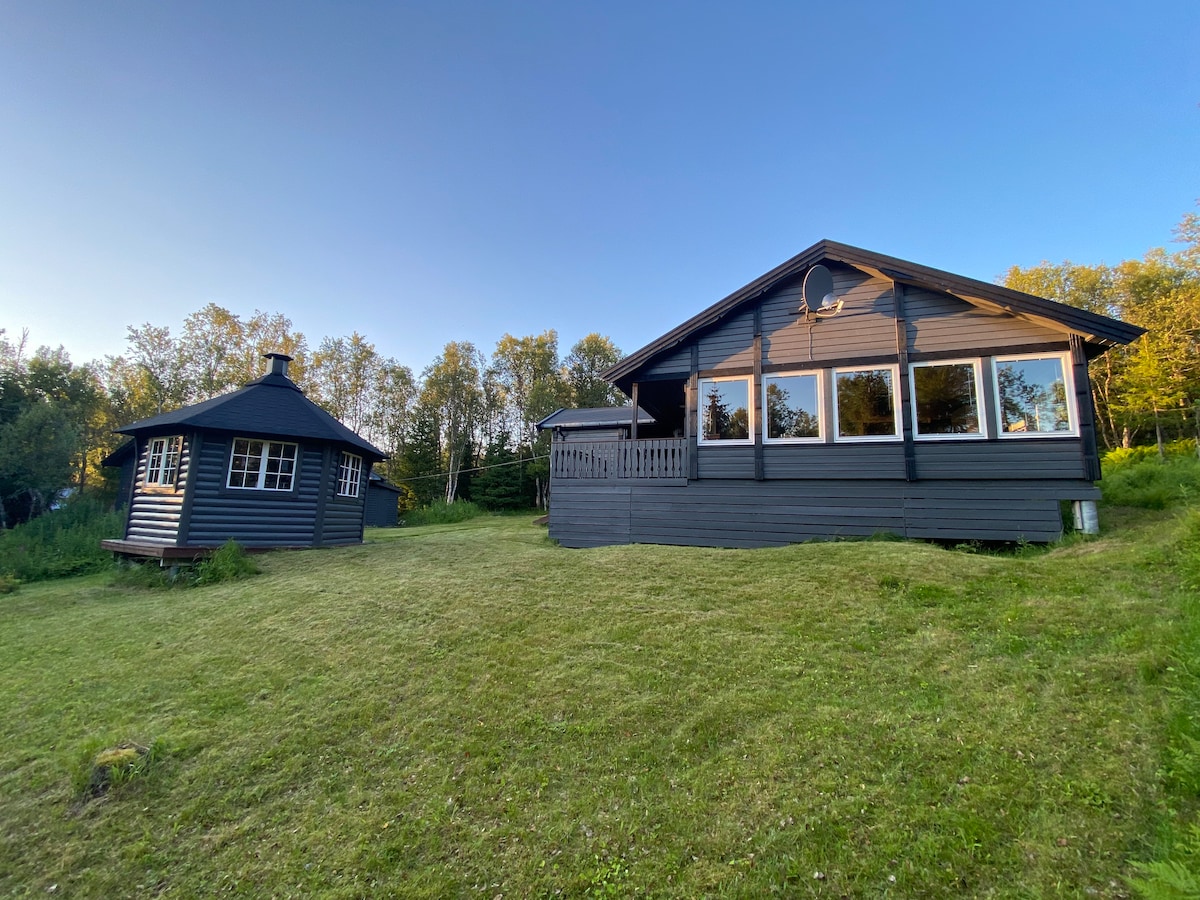 A spacious cabin is surrounded by lush greenery, featuring large windows that allow natural light to illuminate the interior. Beside it, a smaller structure with a circular design adds to the rustic charm of the setting. The area is marked by well-maintained grass.