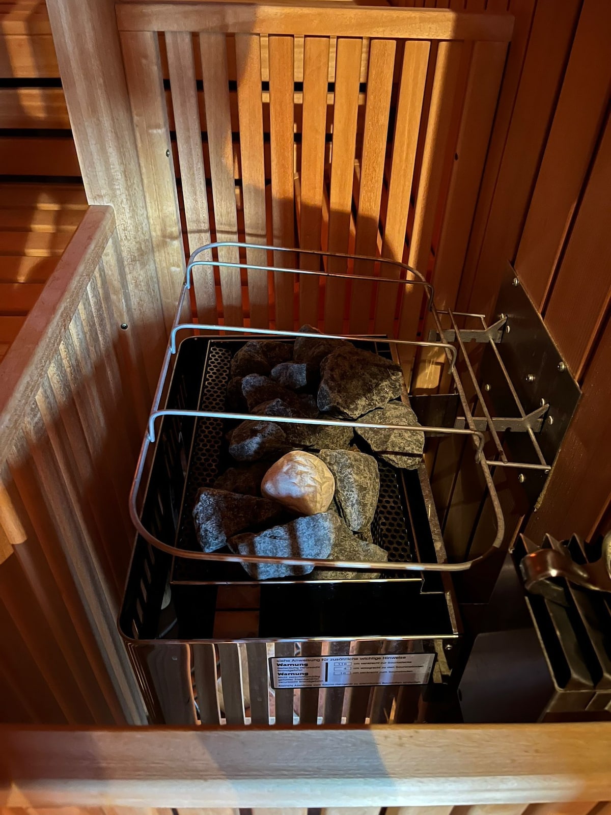 A sauna heater is shown with a metal rack filled with smooth stones, ready for use. The wooden panels of the sauna create a warm ambiance, with shadows playing across the surface. Metal fixtures are visible, indicating the sauna's functionality.