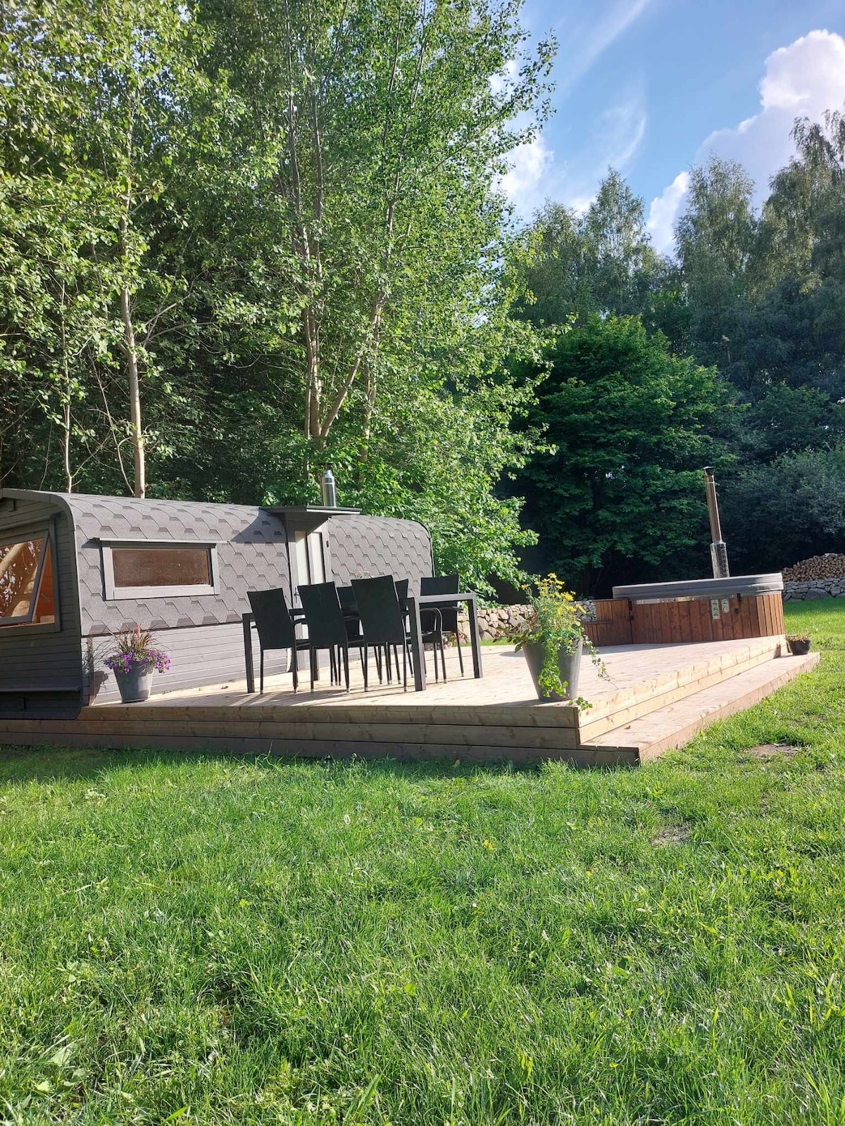 A rustic cabin is nestled among trees, featuring a wooden deck with a dining area set for six. A hot tub is positioned nearby, surrounded by greenery and potted flowers, creating a serene outdoor retreat. Sunlight filters through the leaves, casting a natural ambiance.