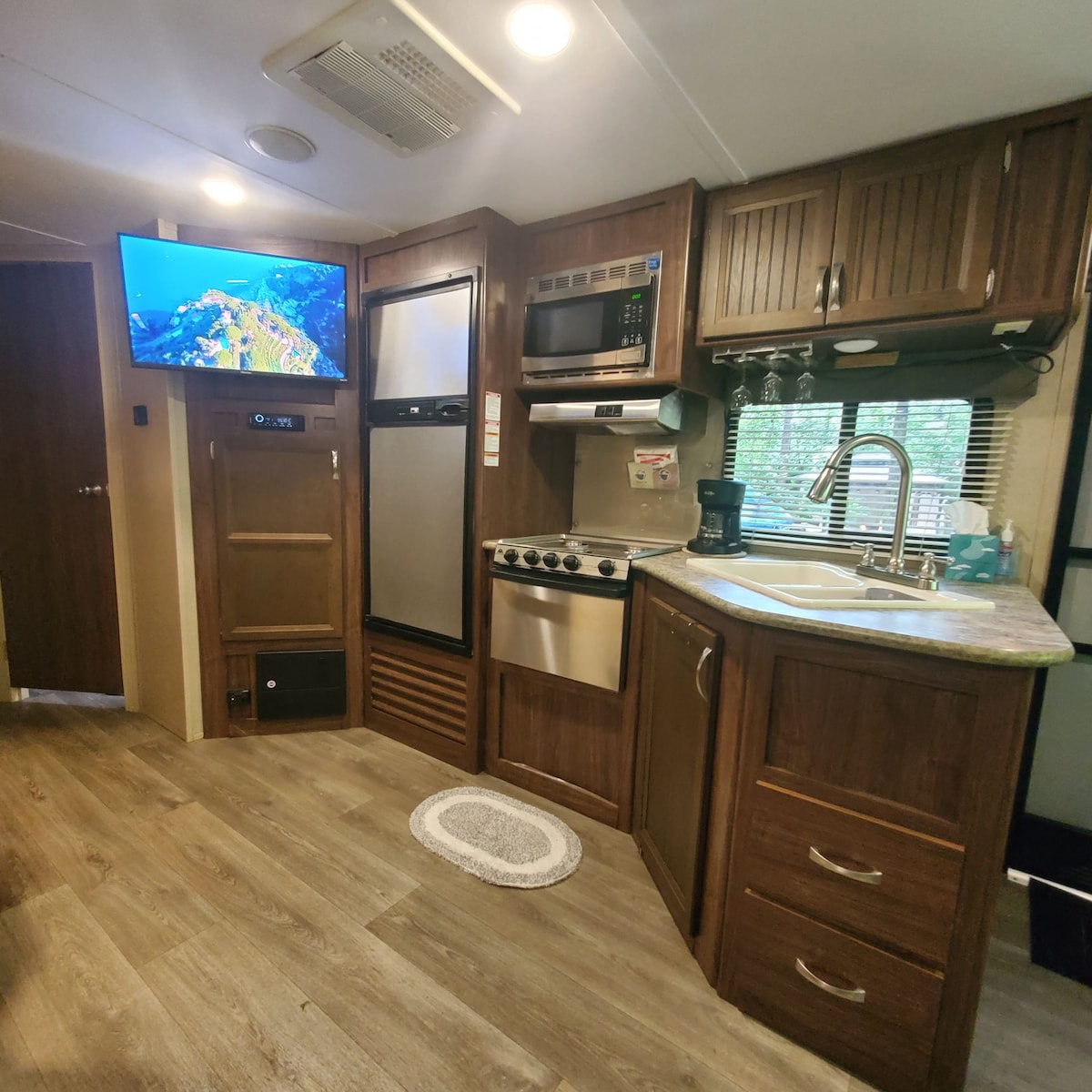 The kitchenette is equipped with modern appliances, including a refrigerator, stove, and microwave. A flat-screen TV is mounted on the wall, and a sink is situated next to a window, allowing natural light to enter the space. Wood cabinetry adds a warm touch.