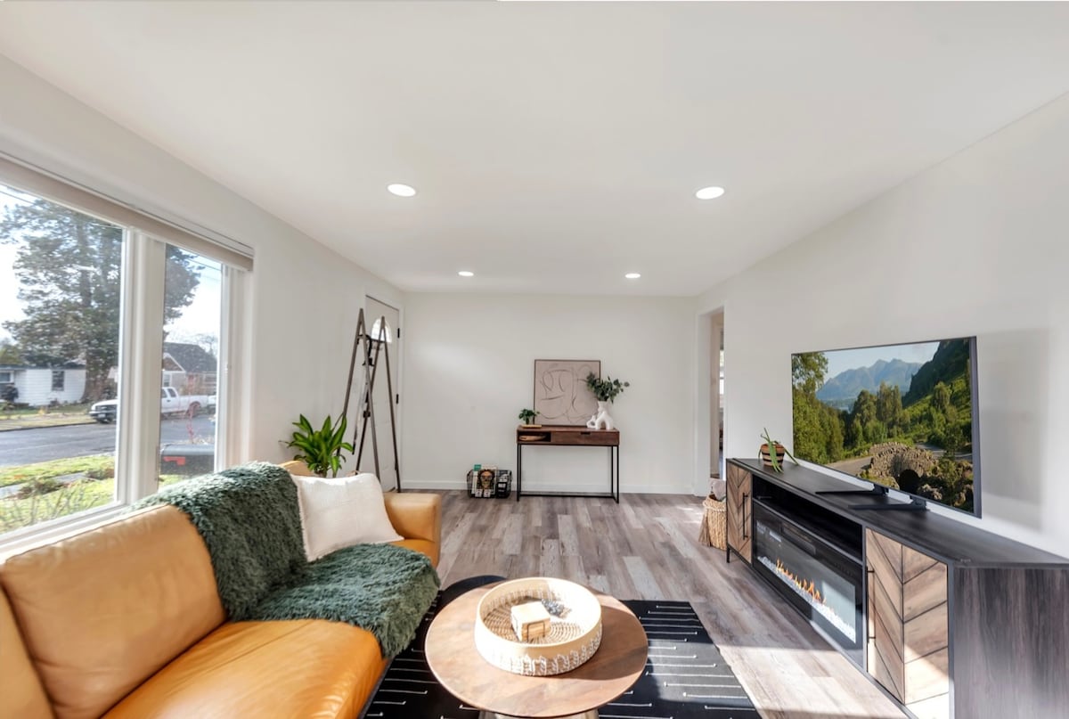 A bright living area is highlighted by a large leather sofa adorned with a soft throw blanket. A round coffee table sits at the center, accompanied by a console with a television displaying scenic natural landscapes. Large windows allow natural light to fill the space.