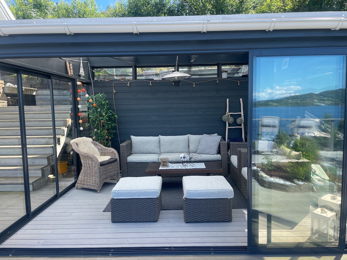 A spacious enclosed terrace features comfortable seating arrangements, including a gray seating area with plush cushions and a central coffee table. Natural light filters through large glass panels, providing views of the surrounding landscape. Steps leading to an outdoor area can be seen in the background.