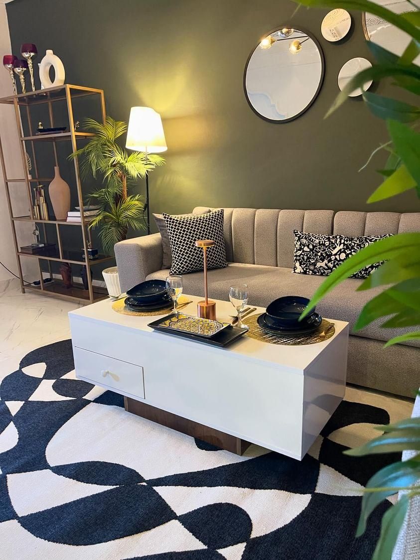 A stylish living area features a gray sofa adorned with patterned cushions, positioned against a dark green wall. A white coffee table is set with dinnerware and glasses, complemented by a decorative tray. A floor lamp and a plant add warmth to the space.