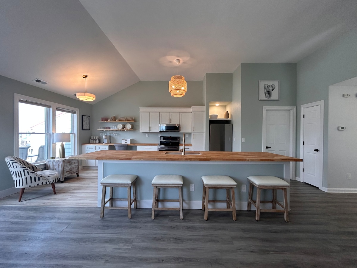 An open-concept kitchen and living area showcase a spacious wooden countertop with four stools positioned at the base. Light walls and a vaulted ceiling create an airy environment, while large windows allow natural light to fill the space. Contemporary furnishings add to the welcoming atmosphere.