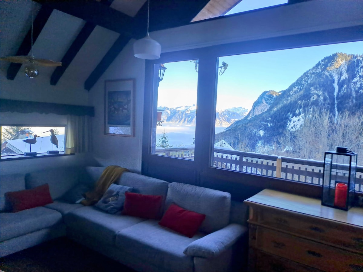 A spacious living area features a comfortable sectional sofa adorned with red cushions, positioned near large windows. Natural light floods the space, revealing stunning views of the mountains and valley. Decorative art pieces add character to the room, enhancing its cozy atmosphere.