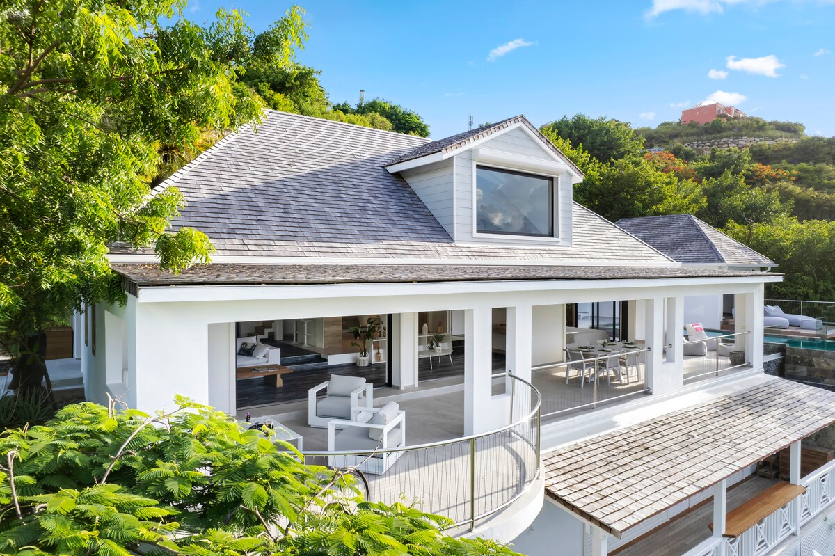 The exterior of the villa is displayed, highlighting a spacious terrace that wraps around the building. Large glass doors provide a view of the inviting indoor living area, while lush green trees surround the property, emphasizing the natural setting.