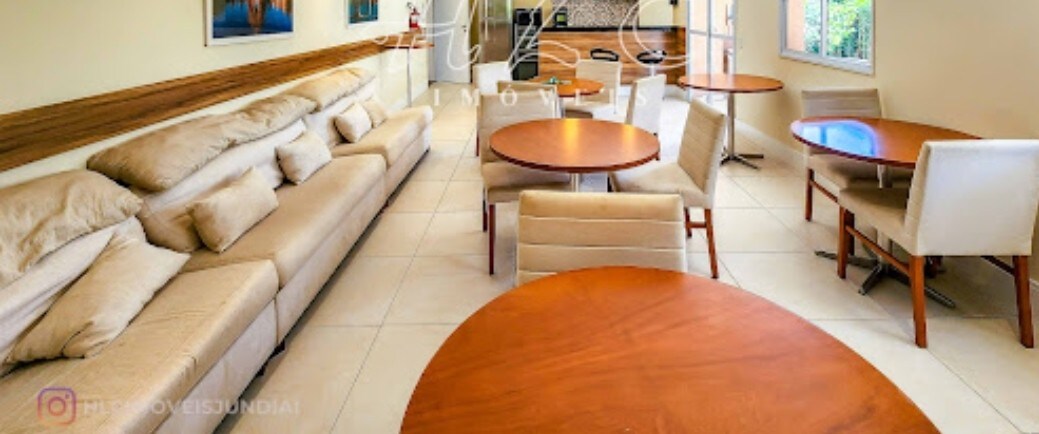 A spacious common area is presented with light-colored sofas and multiple round dining tables. Soft cushions adorn the seating, while natural light filters through large windows, offering a view of the outdoor greenery.