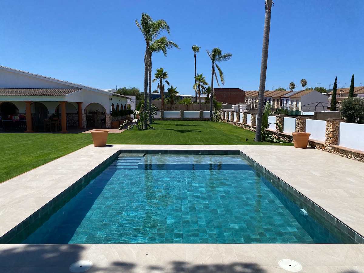 A refreshing swimming pool is centered within a spacious outdoor area, surrounded by well-maintained grass. Palm trees sway gently in the background, and a patio section is visible to one side, providing a relaxed environment for families.