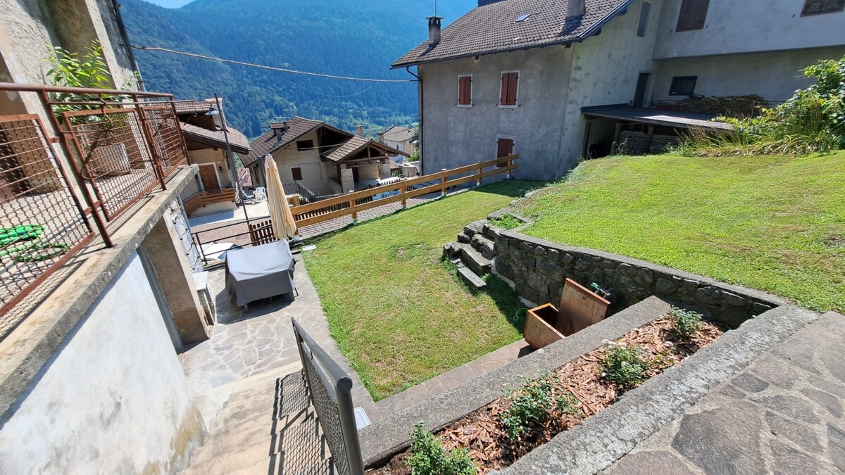 The garden area features a neatly cut grassy space surrounded by stone pathways and low stone walls. Raised garden beds are visible, along with a few steps leading to the entrance of the flat. The scenic mountains are visible in the background, enhancing the outdoor view.