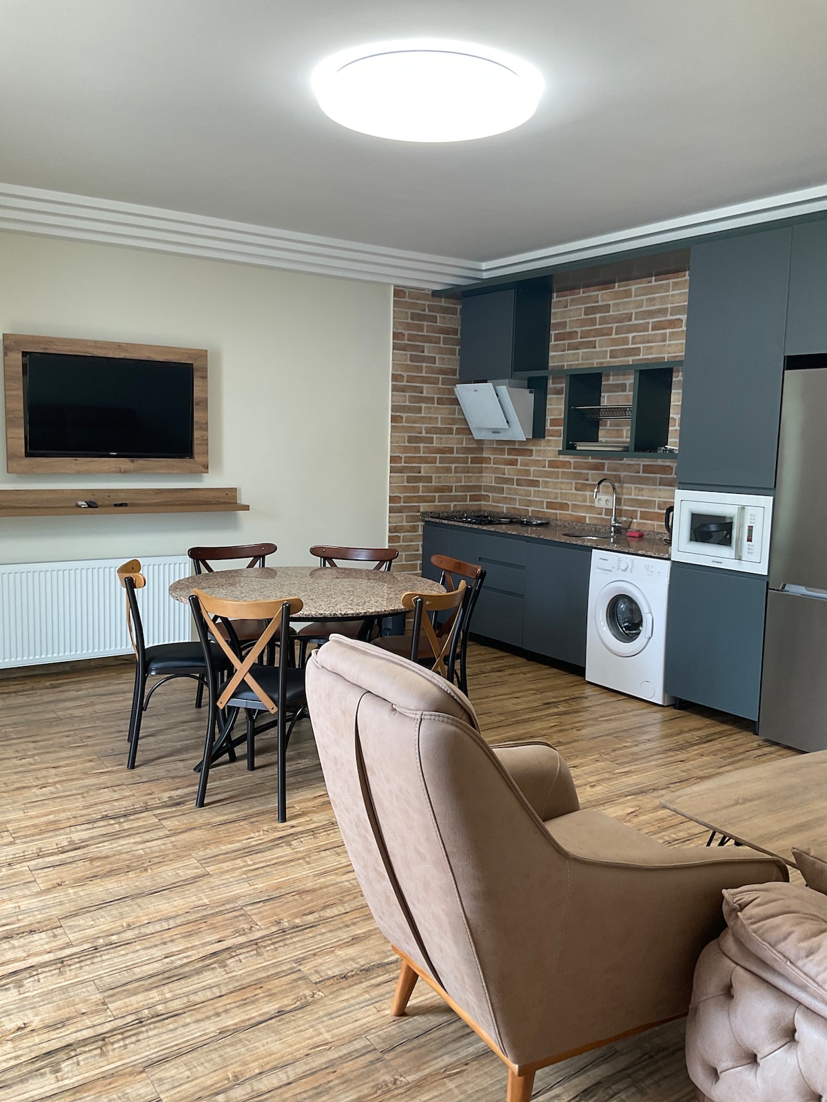 The open living area features a dining table with six chairs, a comfortable armchair, and a mounted flat-screen TV. The kitchen includes modern appliances and a brick accent wall, while a washing machine is neatly positioned near the dining space.