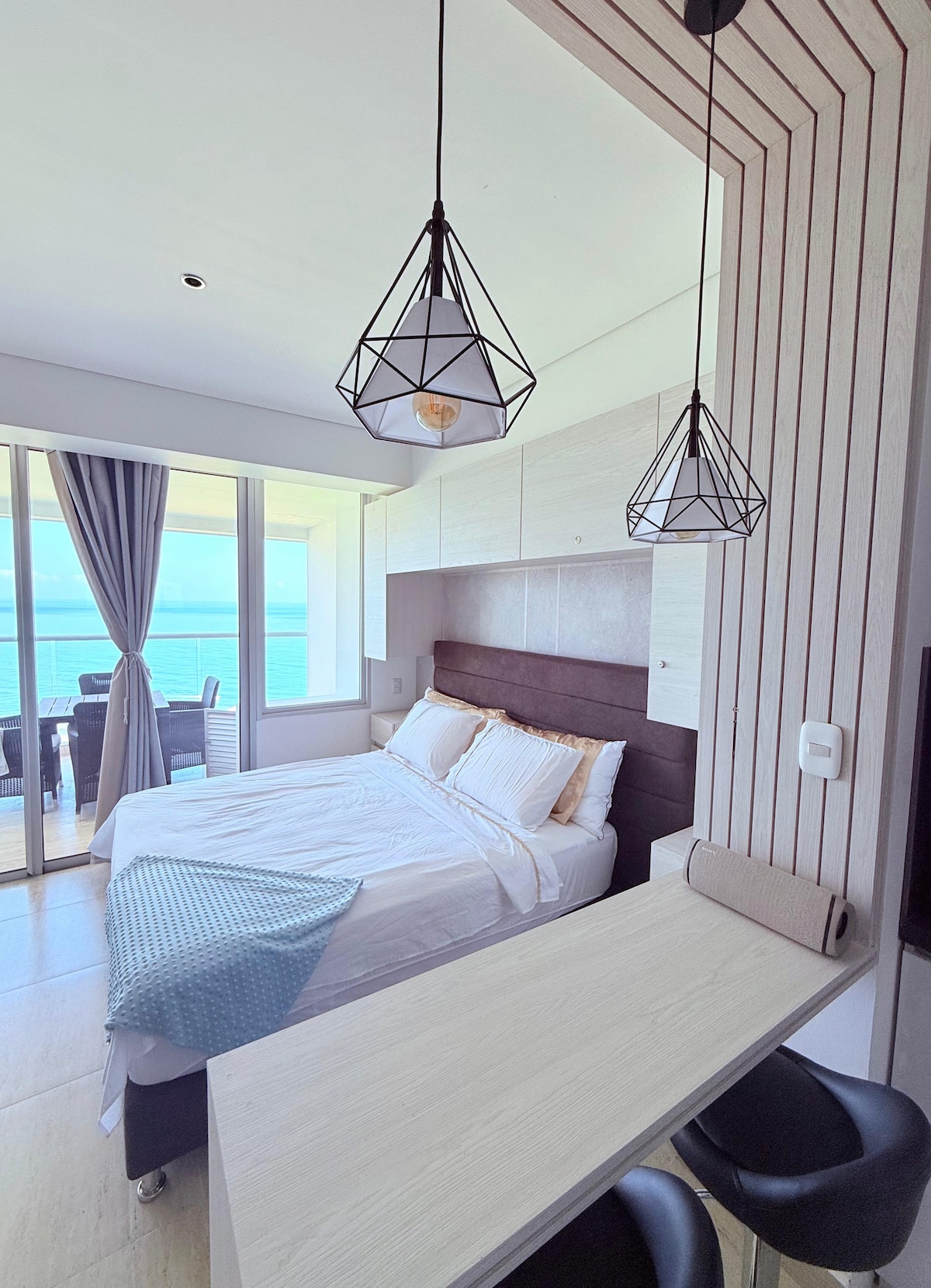 A compact bedroom is presented with a spacious bed featuring crisp white linens. Large windows allow natural light and provide views of the ocean. Two pendant lights hang above a small dining area with two black chairs and a light wood table.