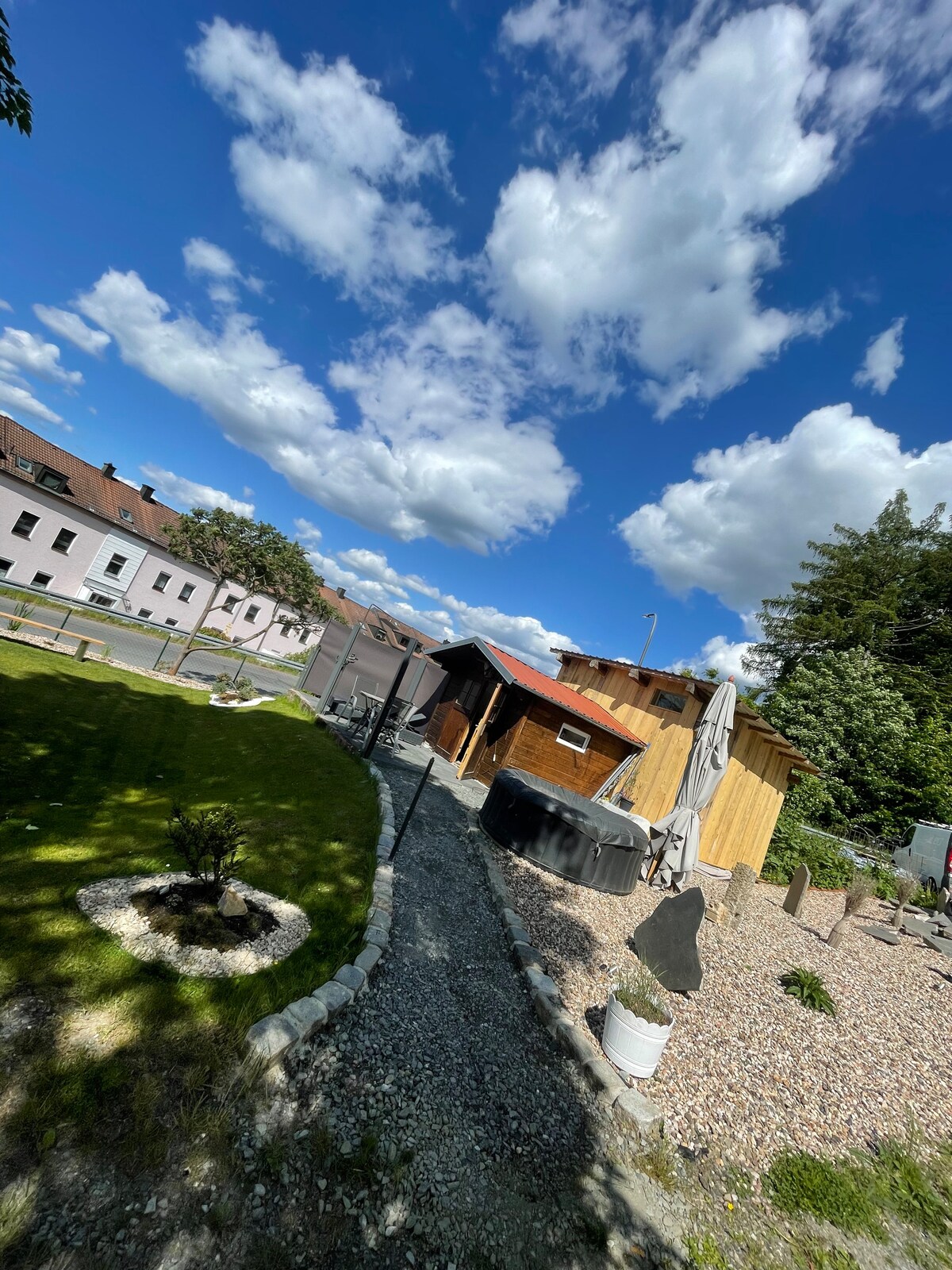 A well-maintained outdoor space surrounds the tiny house, featuring a mix of gravel and grass pathways. A circular garden bed with a small plant is visible, along with a covered seating area. The sky is adorned with fluffy clouds, complementing the serene environment.