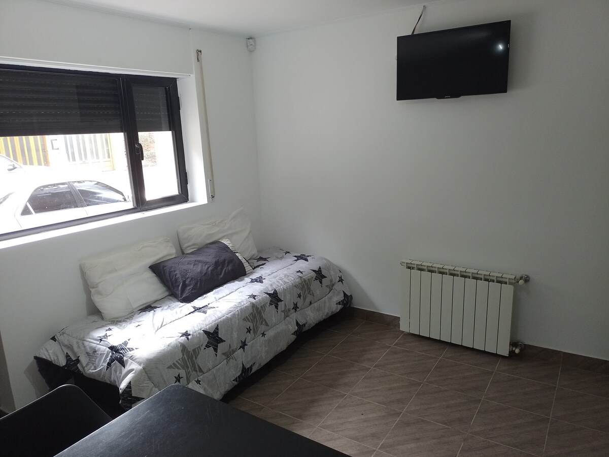 A bright sleeping area features a space-saving bed adorned with a star-patterned duvet cover. A flat-screen television is mounted on the wall, and a large window offers a view of the outside. Radiant heat is provided by a wall-mounted heater.