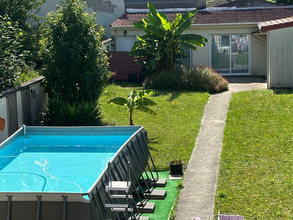A clear blue pool is positioned within a grassy area, accompanied by lounge chairs arranged along its edge. Lush greenery and tropical plants enhance the setting. A well-defined pathway leads to a cozy dwelling with expansive glass doors, suggesting easy access to indoor spaces.