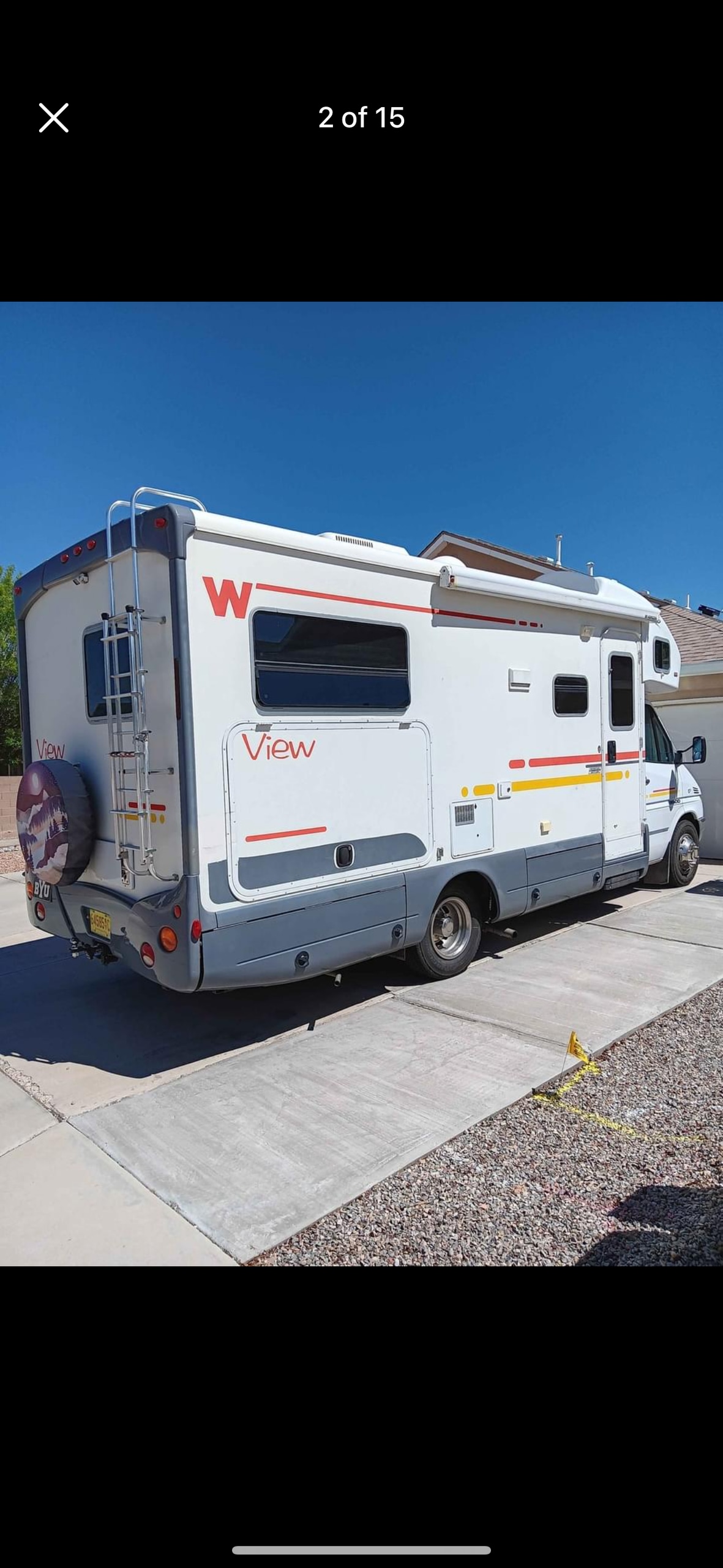 A motor home is parked on a paved surface, featuring a streamlined design and sizeable rear window. Distinctive markings adorn the vehicle's side, while the clear blue sky looms overhead, emphasizing its outdoor setting. The surroundings are tranquil and inviting, suitable for relaxation.