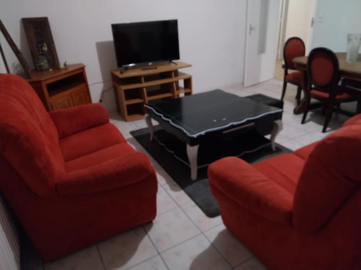 A living area is presented with two red armchairs facing a black coffee table, positioned on a dark carpet. A television is mounted on the wall, while a wooden cabinet and dining table are visible in the background.