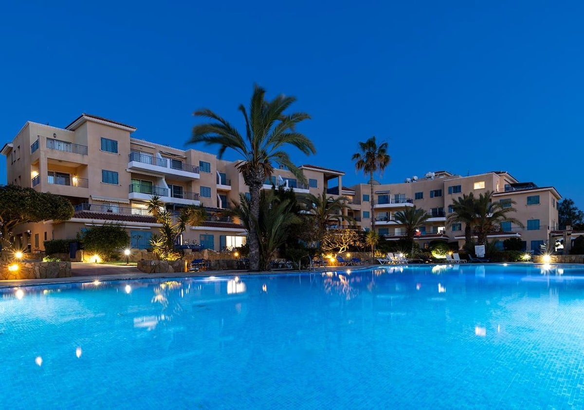 An illuminated pool reflects the evening sky, surrounded by palm trees and well-maintained landscaping. The nearby buildings are softly lit, enhancing the serene environment. The scene showcases a harmonious blend of natural and architectural elements, creating a peaceful retreat for guests.