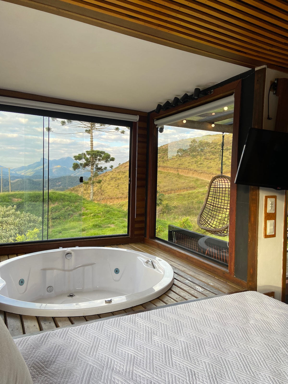 A bright space showcases a round hot tub positioned near large windows with expansive views of the surrounding hills. Wooden accents frame the area, while a hanging chair offers a cozy spot to relax. A comfortable bed is visible in the foreground.