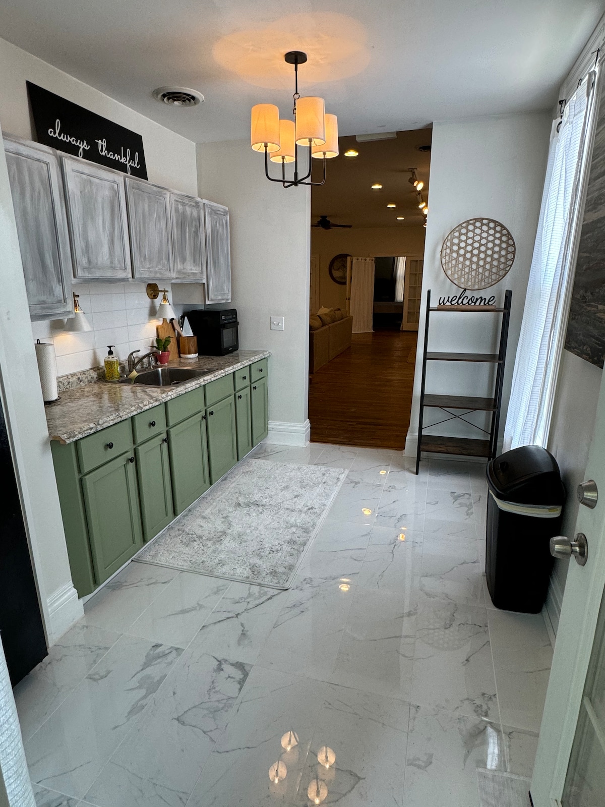 A compact kitchen space is fitted with green cabinetry and a gray countertop. Essential appliances, including a coffee maker and microwave, are positioned along the countertops. Lighting is provided by a modern chandelier, and a welcome sign is visible near the entrance.
