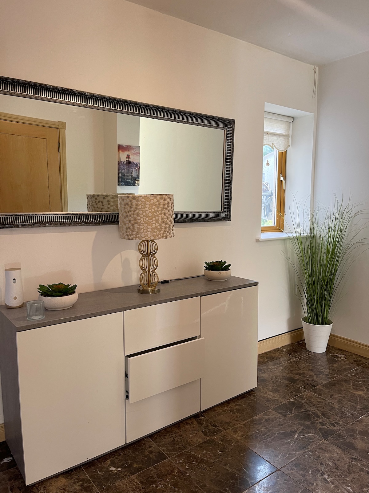 A light-filled entryway features a sleek white dresser topped with a decorative lamp and small plants. A large mirror is mounted above, reflecting the room's openness. A window allows natural light to flow in, with a tall indoor plant in a white pot positioned nearby.