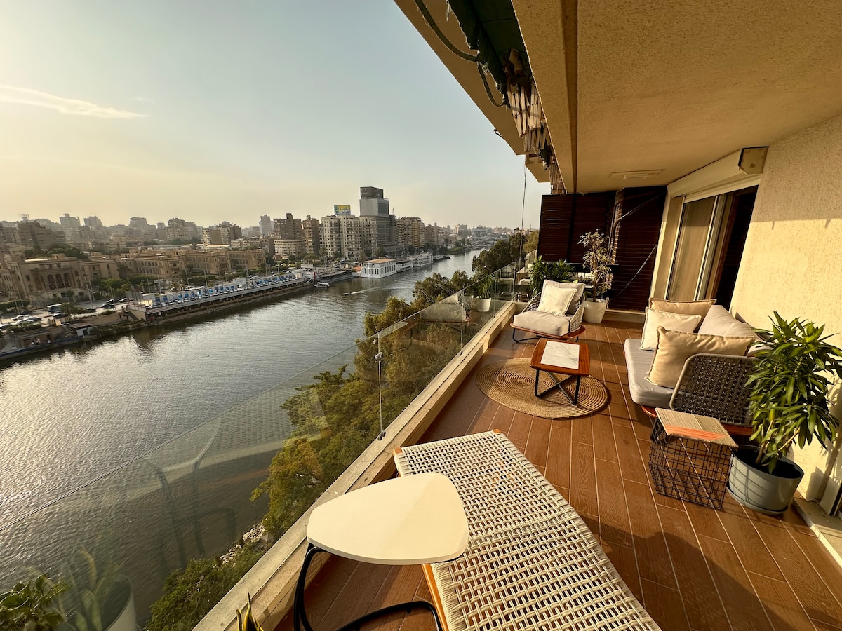 A serene balcony area is captured, featuring comfortable seating on wooden decking. A small table is positioned beside a stylish chair, overlooking the tranquil Nile river and bustling cityscape beyond. Lush greenery enhances the inviting outdoor space, complemented by the warm glow of the setting sun.