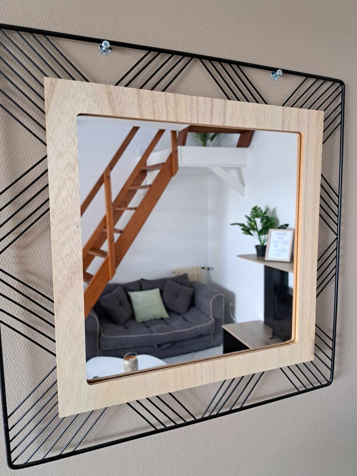 A framed mirror is mounted on the wall, reflecting a cozy living area featuring a gray sofa with a green cushion. A staircase leading to a mezzanine is visible in the background, and a plant is placed on the shelf by the fireplace.