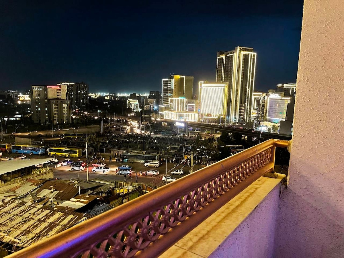 A balcony view showcases a lively nighttime scene of Addis Ababa, with brightly lit skyscrapers illuminating the skyline. Streets below pulse with traffic, as the urban landscape comes alive against a dark sky, creating an engaging city atmosphere.