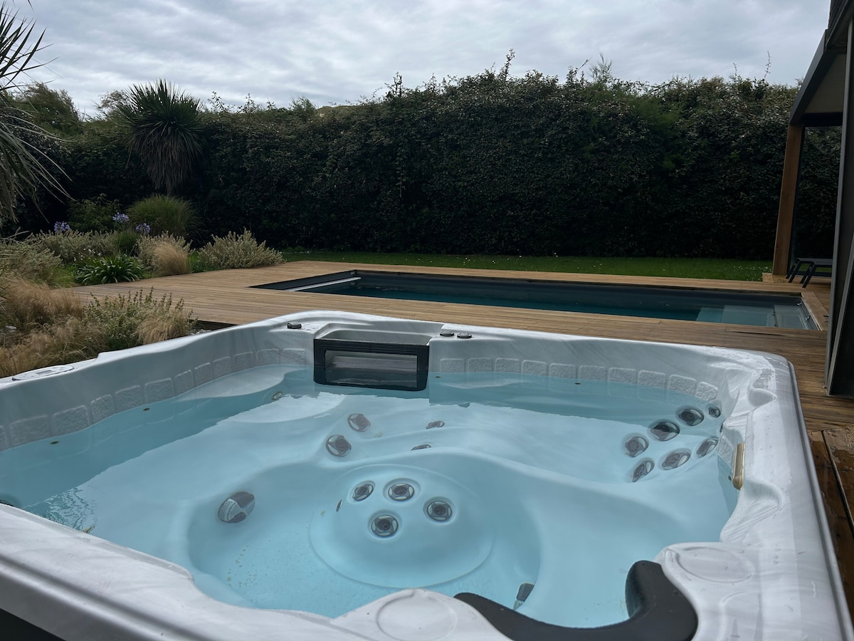 A hot tub is shown, surrounded by greenery and wooden decking. In the background, a swimming pool is visible, bordered by grass and plants. Overcast skies are present, creating a serene environment.