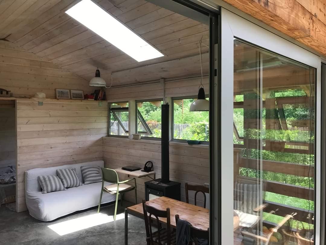 An inviting common space features large windows allowing natural light to fill the area. A cozy couch with striped cushions is positioned alongside a wooden dining table. A small black stove is placed near the seating, and a workspace is situated within the room.