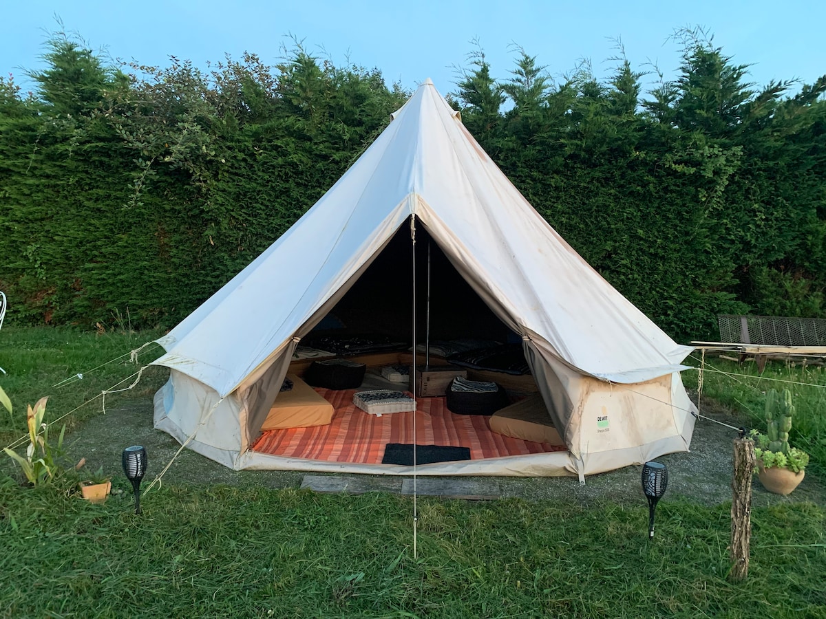 A tent with a conical shape stands in a grassy area, surrounded by greenery. The entrance is open, revealing a cozy interior with several bedding areas, including mats and cushions arranged on a checkered floor covering. Subtle outdoor lighting fixtures are positioned nearby.