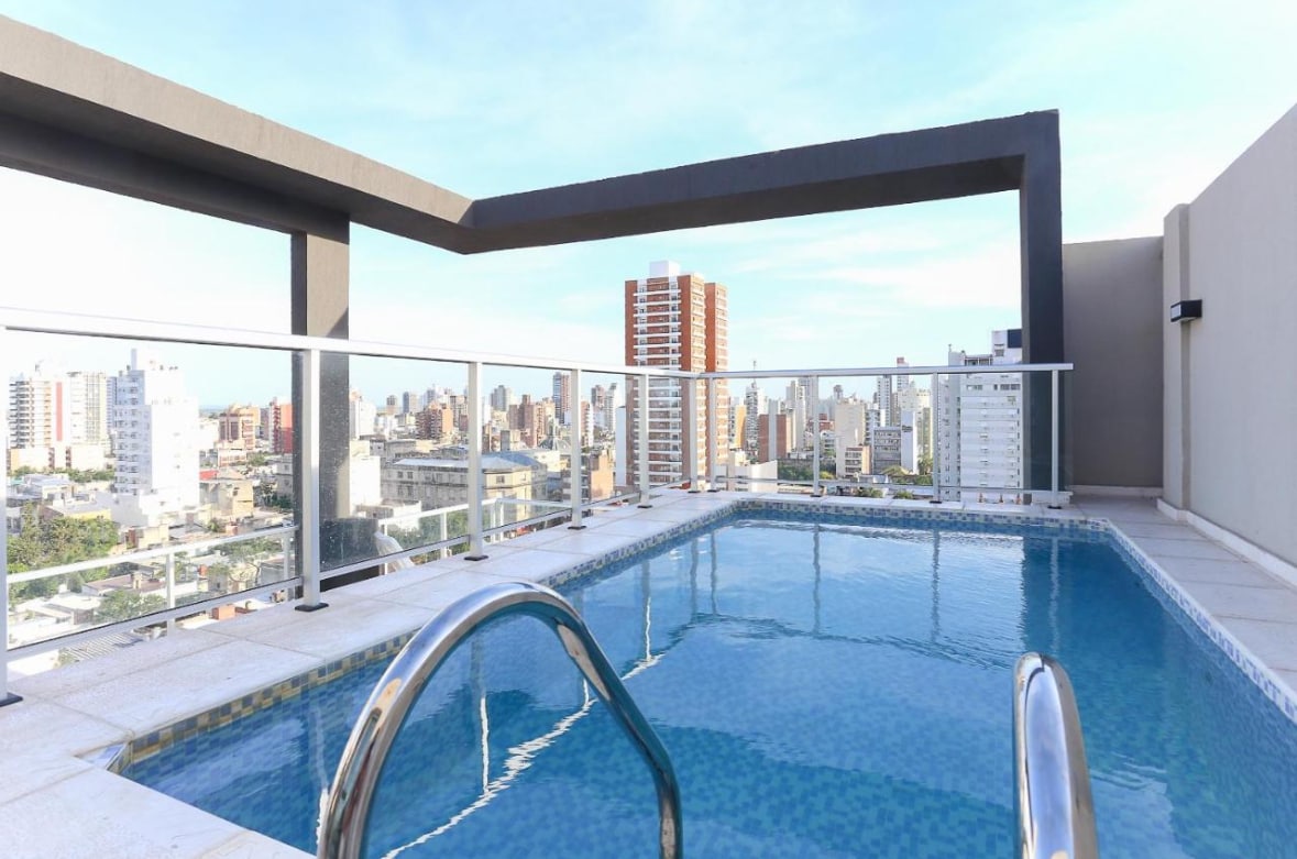 An inviting rooftop pool with a clear blue surface is framed by sleek glass railings, offering panoramic views of the city skyline. The surrounding area features modern buildings and a bright sky, providing a serene backdrop for relaxation.