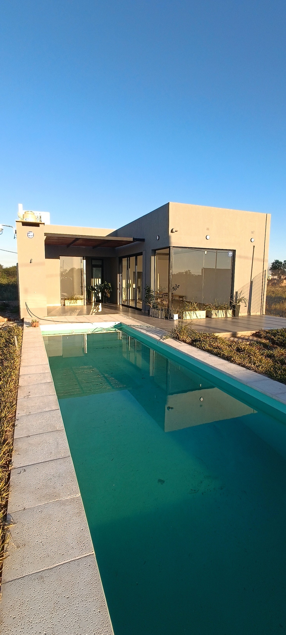 A modern single-story building features large glass panels that provide natural light and views of the surrounding landscape. A sleek, clear pool runs parallel to the structure, reflecting the blue sky above, while minimal landscaping frames the area.