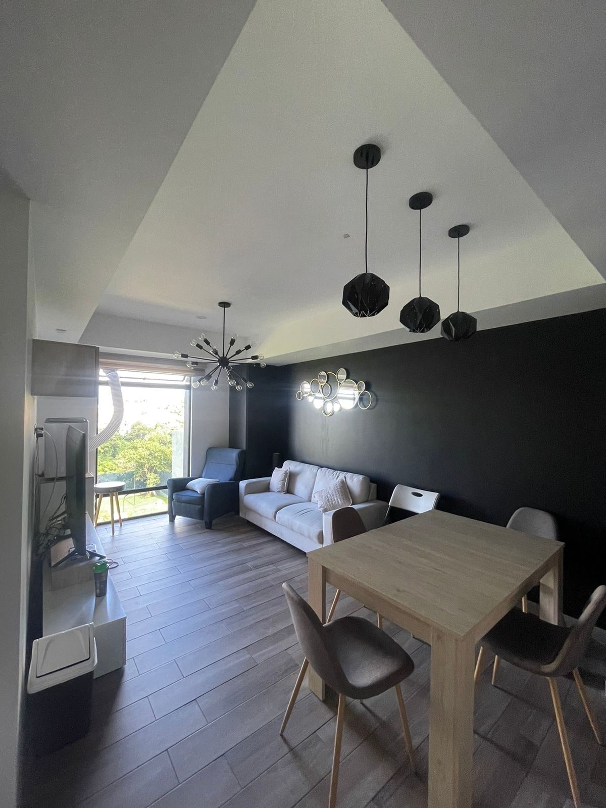 A well-lit living space features a comfortable sofa against a bold black wall. A modern dining table with four chairs is positioned nearby. Unique light fixtures hang from the ceiling, while a large window allows natural light to illuminate the room.