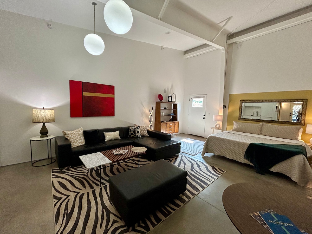 An open-concept living space features a modern black sofa and a coffee table on a striking zebra-patterned rug. A spacious bed is positioned adjacent to a decorative mirror. Soft lighting is provided by pendant lights and two bedside lamps, creating a calming ambiance.
