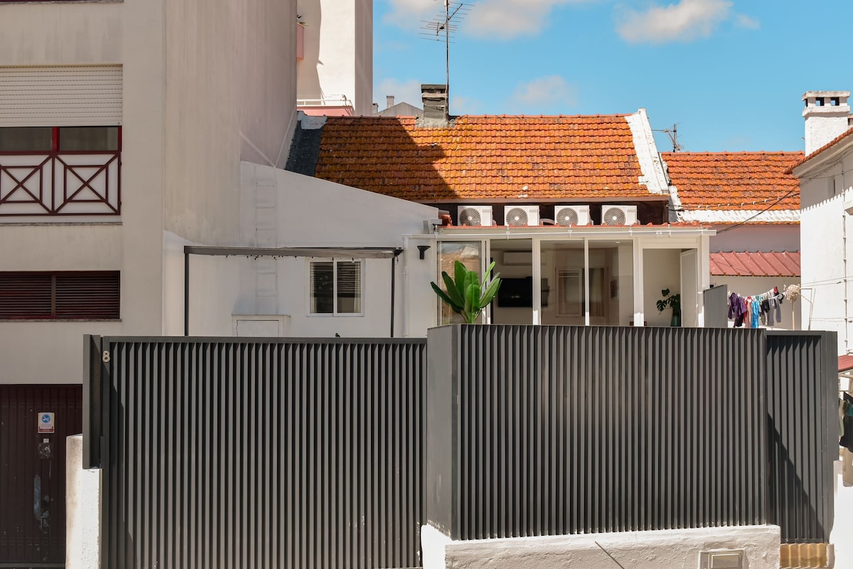 A modern black metal fence provides privacy to a residence featuring a tiled roof. A balcony is visible on the upper level, with laundry hanging outside. The surrounding area includes light-colored buildings and clear blue sky, creating a peaceful urban setting.