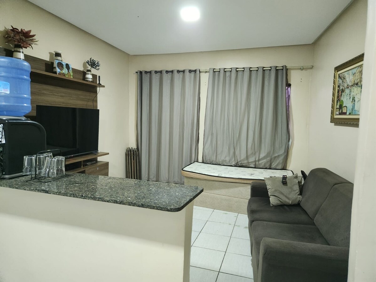 An open-plan living area features a modern kitchenette with a granite countertop. A comfortable gray sofa is positioned against the wall, complemented by a large window covered with light gray curtains. A TV unit is mounted nearby, adding to the functionality of the space.