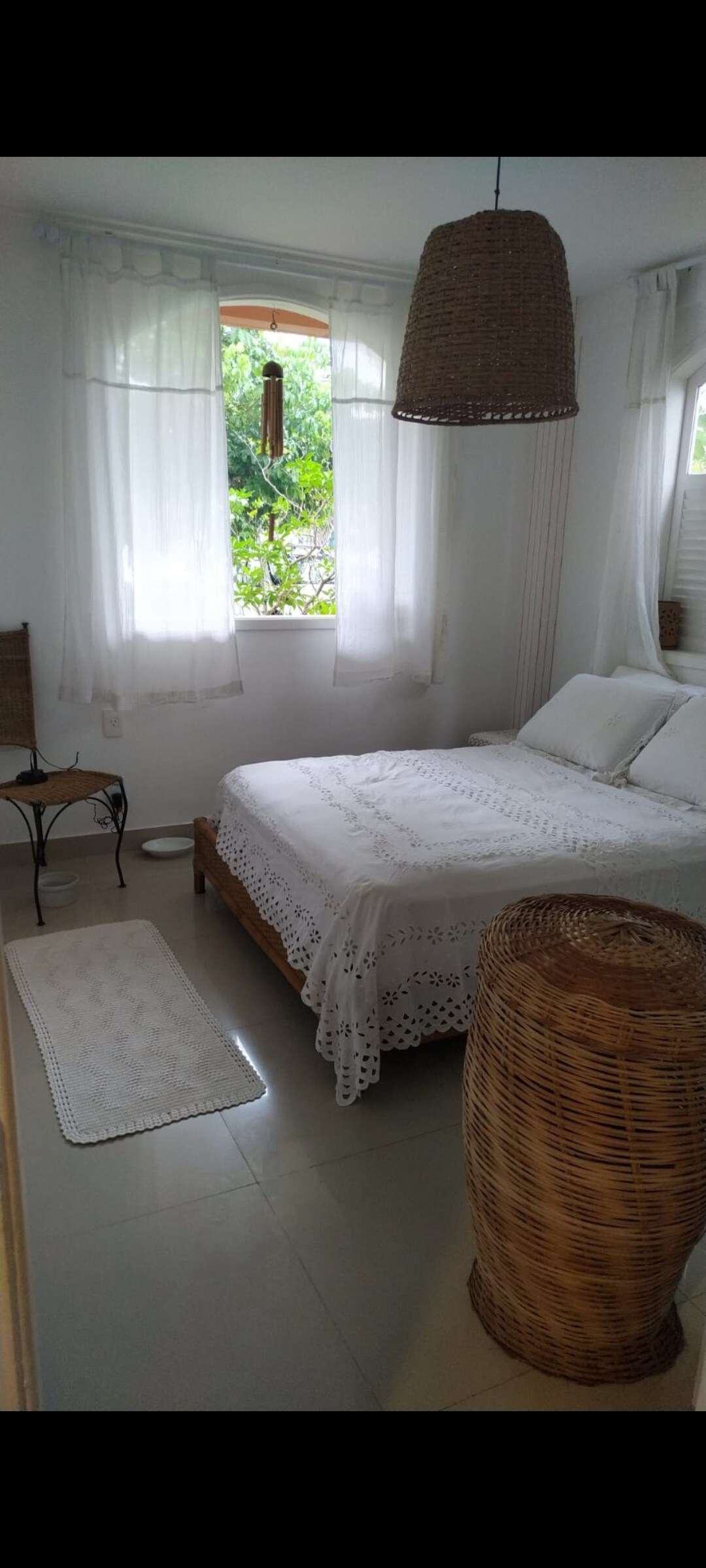 A bright and airy sleeping area features a spacious bed dressed in light linens. Natural light filters through sheer curtains, illuminating the room. A woven pendant light hangs overhead, and a small side table is positioned next to the bed, complementing the natural textures throughout.