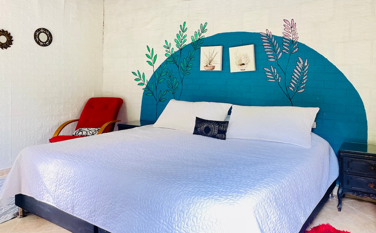 A spacious sleeping area features a large white bed with decorative pillows. A vibrant blue wall is adorned with plant-inspired artwork, enhancing the space. A comfortable red chair is positioned nearby, alongside a dark wooden bedside table.