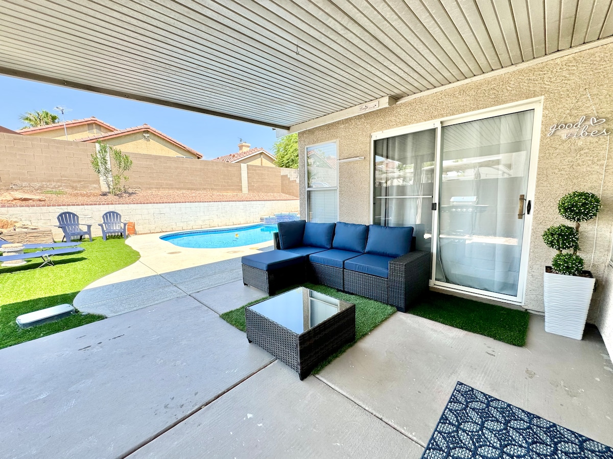 An outdoor seating area features a dark wicker sectional sofa, accompanied by a glass-topped table. A private pool is visible in the background, surrounded by artificial grass. Sun loungers are placed nearby, and a wall enhances the secluded atmosphere.