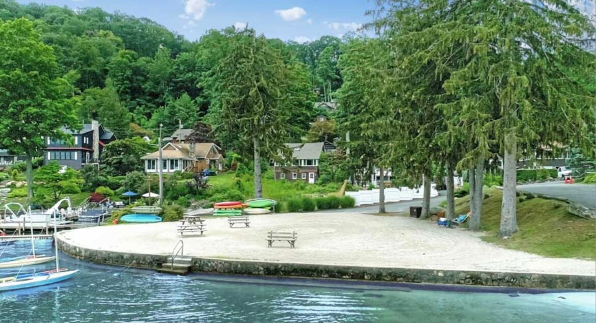 A sandy beach area is framed by lush green trees and surrounded by several colorful boats. Picnic tables are positioned in the vicinity, offering spaces for outdoor gatherings. The background features charming cabins nestled among the trees, enhancing the community atmosphere.