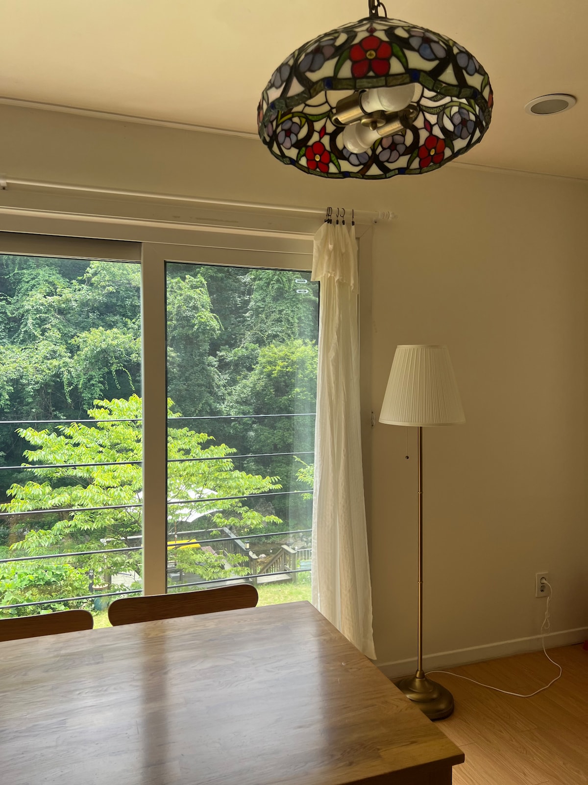 A light-filled room features a wooden dining table beneath a stained glass chandelier. Large glass doors provide a view of lush greenery outside, while a stylish floor lamp stands beside the window, enhancing the space's tranquil ambiance.