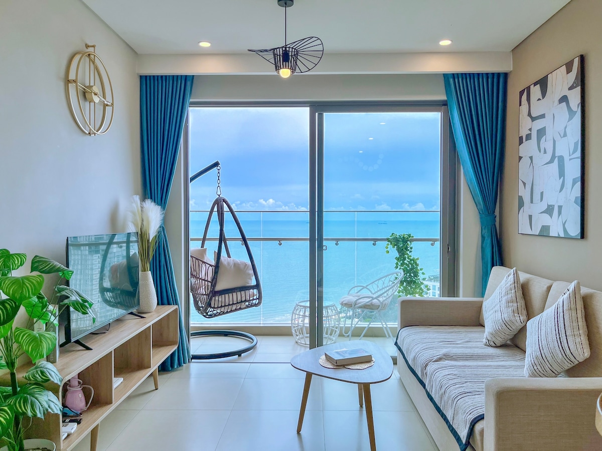 A modern living area features large glass doors opening to a balcony with ocean views. A comfortable sofa is accompanied by a light wooden coffee table. A hanging chair adds an inviting touch, while plants and wall art enhance the space's ambiance.