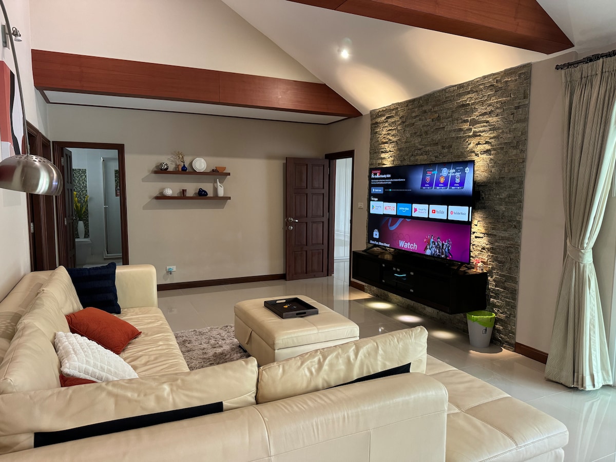The living area features a spacious, light-colored leather sofa adorned with decorative pillows. A large flat-screen television is mounted against a stone accent wall, with additional shelves displaying tasteful decor. Natural light enters through window treatments, creating a comfortable and inviting environment.