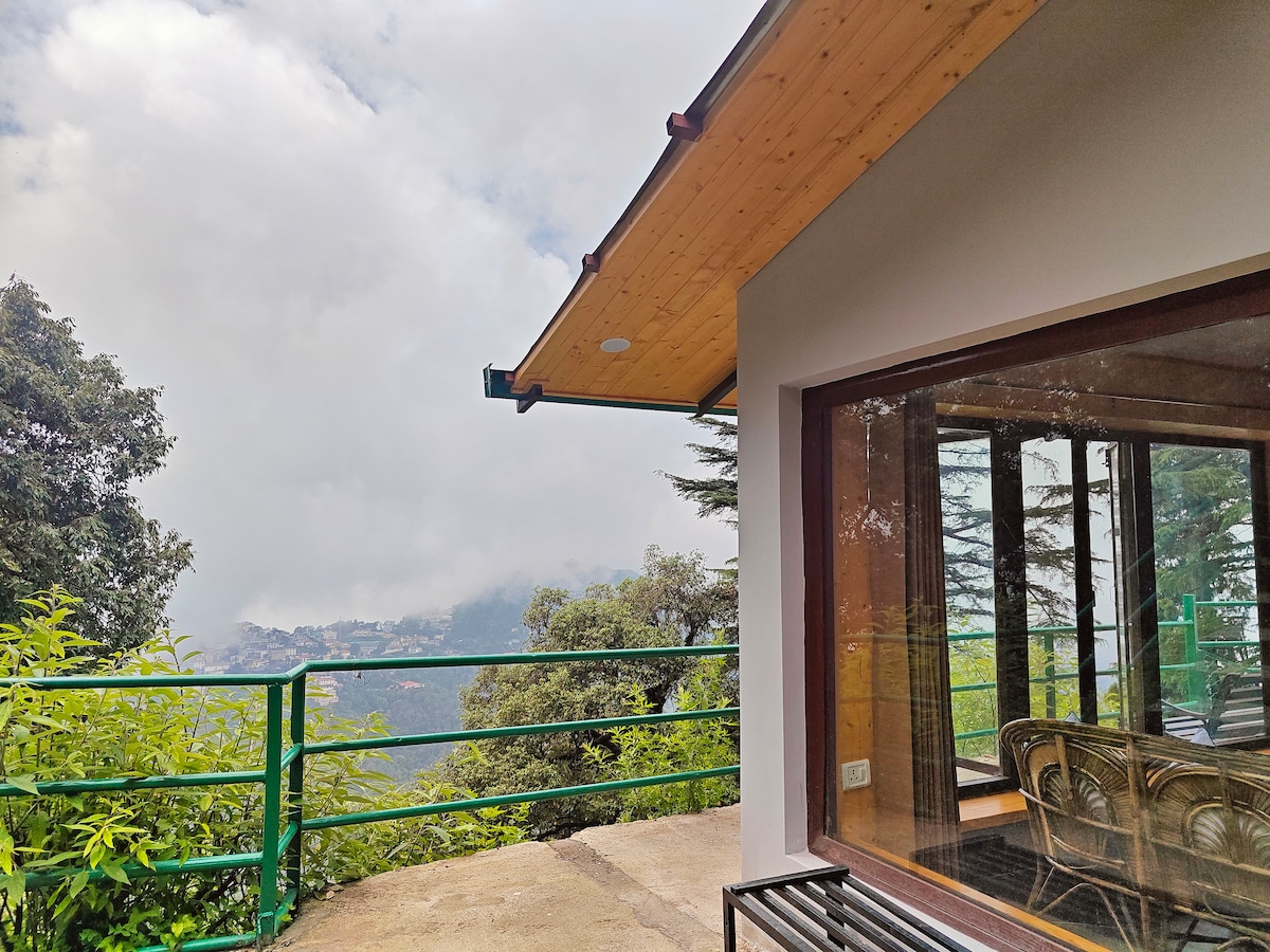 An outdoor deck is visible, featuring a railing that overlooks the valley and mountains. The cabin's wooden roof extends outward, while large glass windows invite natural light into the interior. Lush greenery surrounds the area, blending the space with nature.