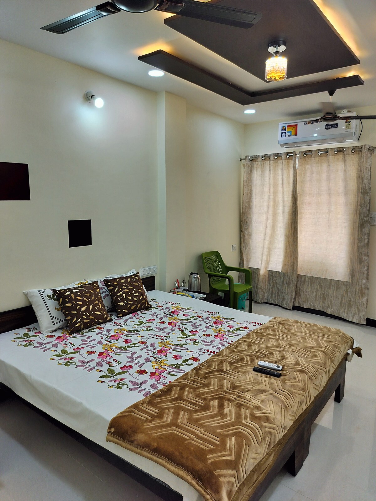A bright sleeping area is equipped with a spacious bed adorned with floral-patterned linens and decorative pillows. A soft blanket is neatly placed at the foot of the bed. A chair sits near the window, which is dressed in light-colored curtains, allowing natural light to enter.