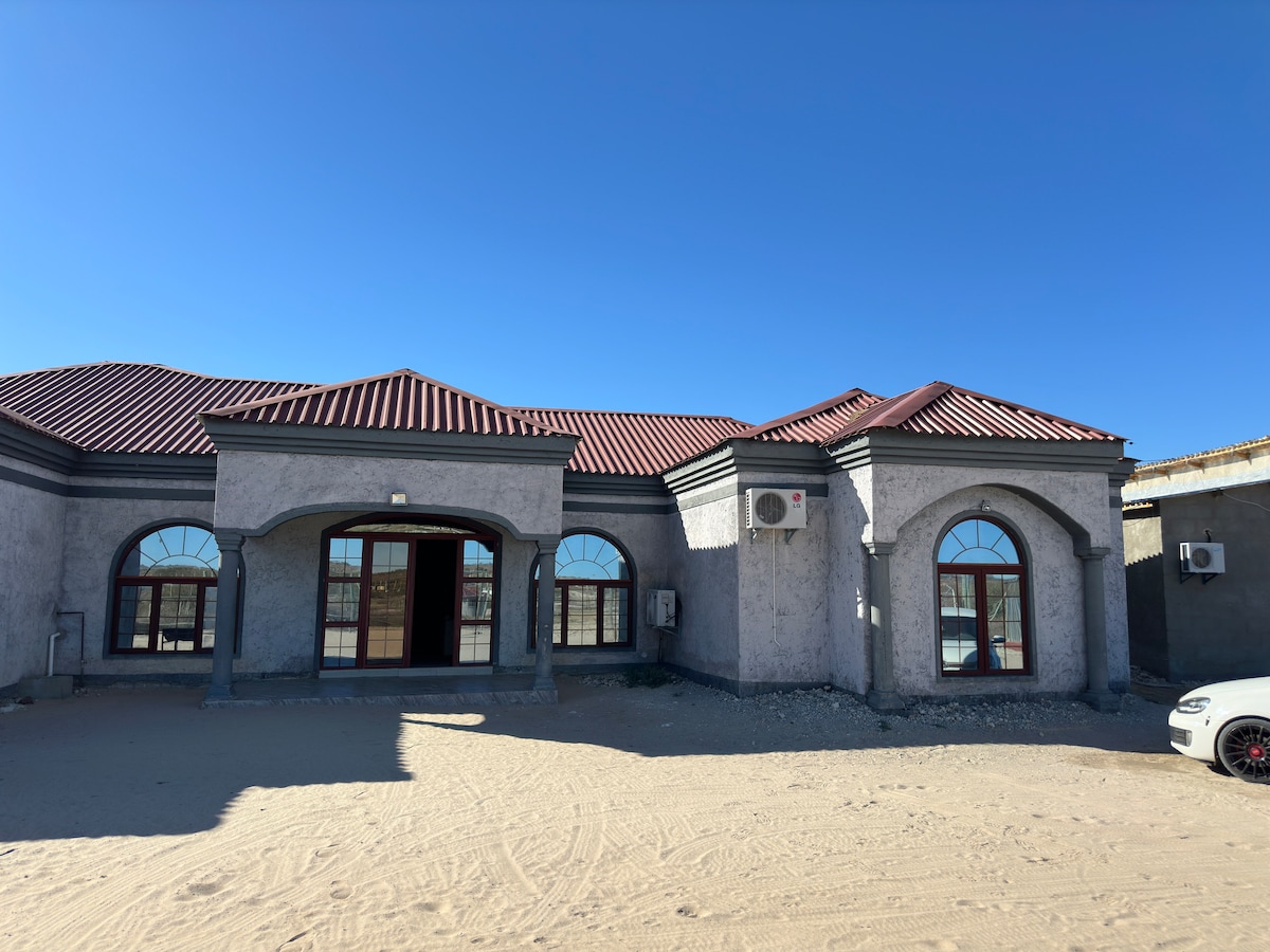 The exterior of the guesthouse features a modern design with a textured facade and red-roofed sections. Arch-shaped windows frame the entrance, allowing natural light to enter. Two air conditioning units are visible on the sides, providing comfort in warm weather.