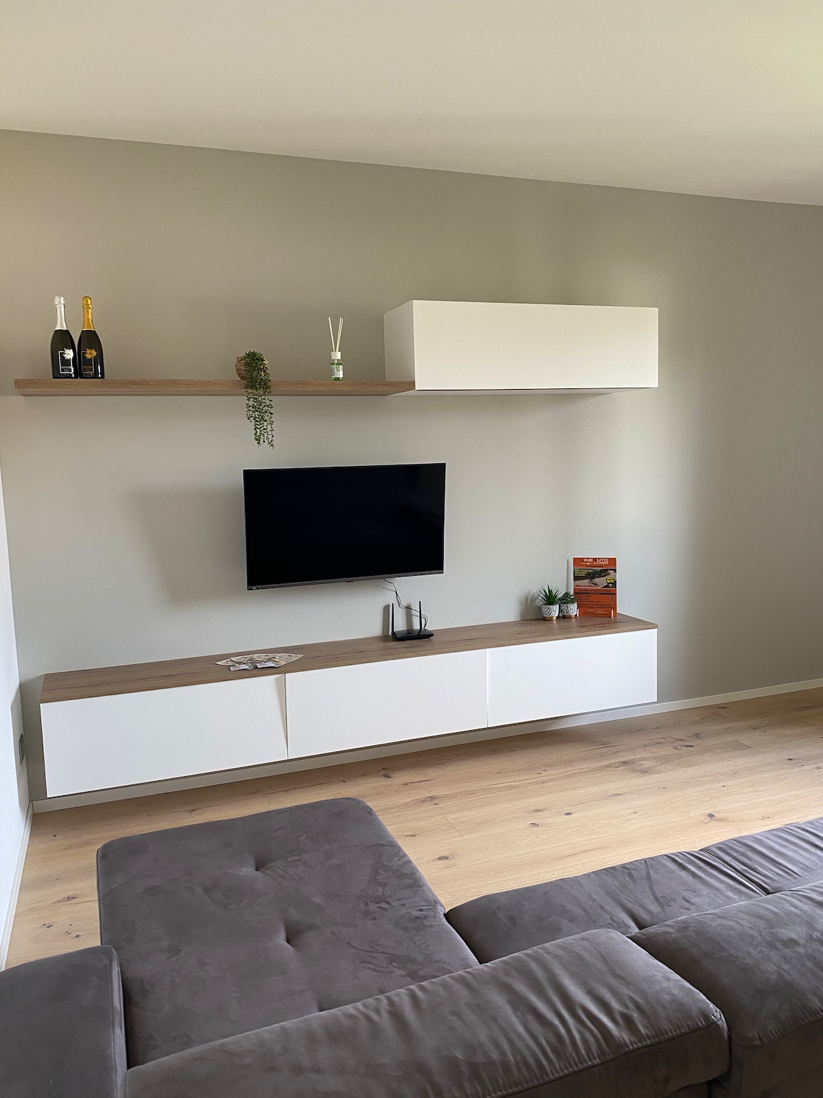 A modern living area features a sleek wall-mounted television above a minimalist console. Light wood shelves hold decorative items, including two bottles and a small plant. A comfortable gray sofa invites relaxation, complementing the soft, neutral-colored walls and light hardwood flooring.