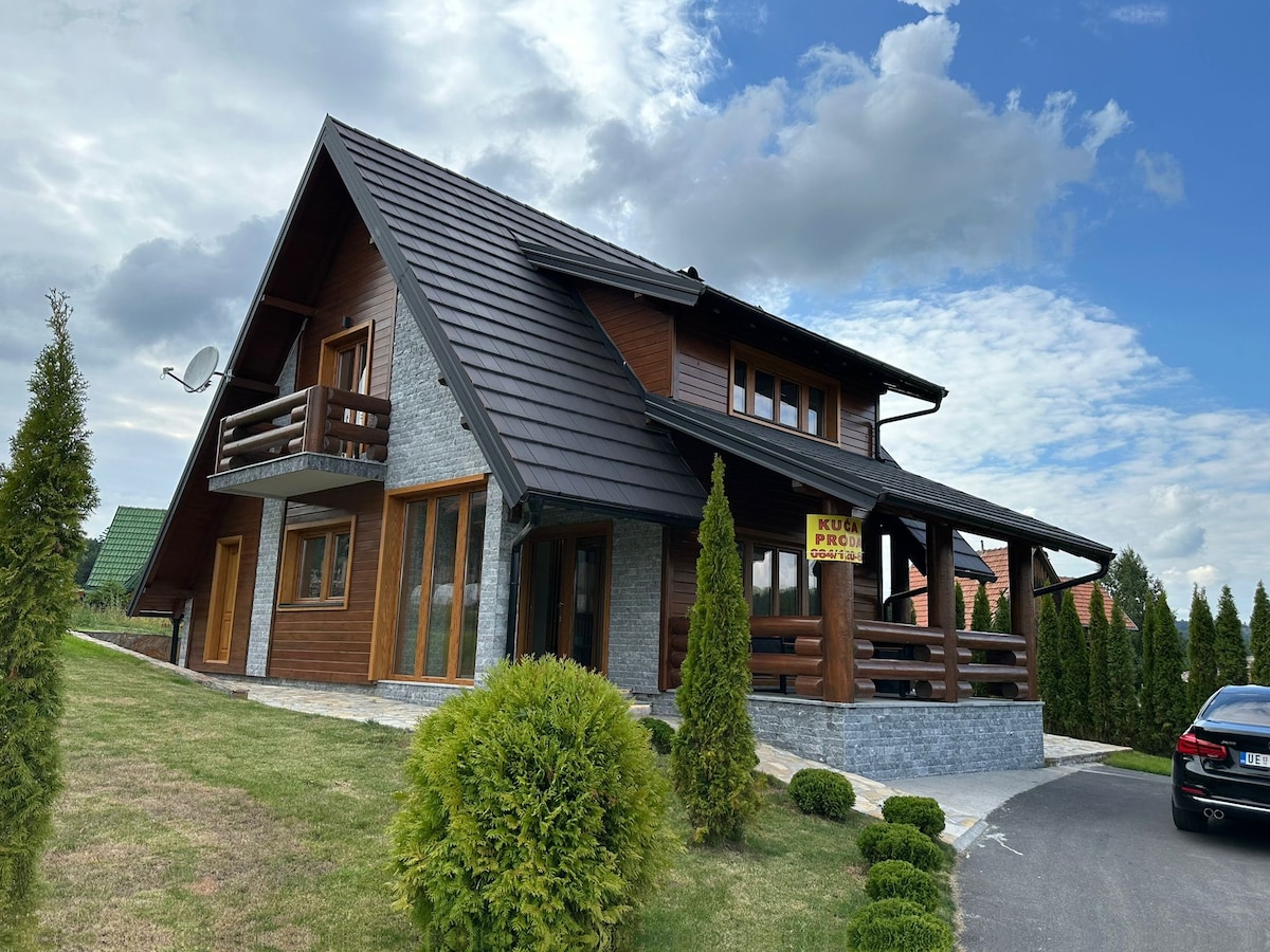 The exterior of the wooden house showcases a steeply pitched roof and a combination of natural wood and stone elements. Lush green landscaping surrounds the property, featuring neatly trimmed shrubs and a wide lawn area, creating a serene and inviting atmosphere.